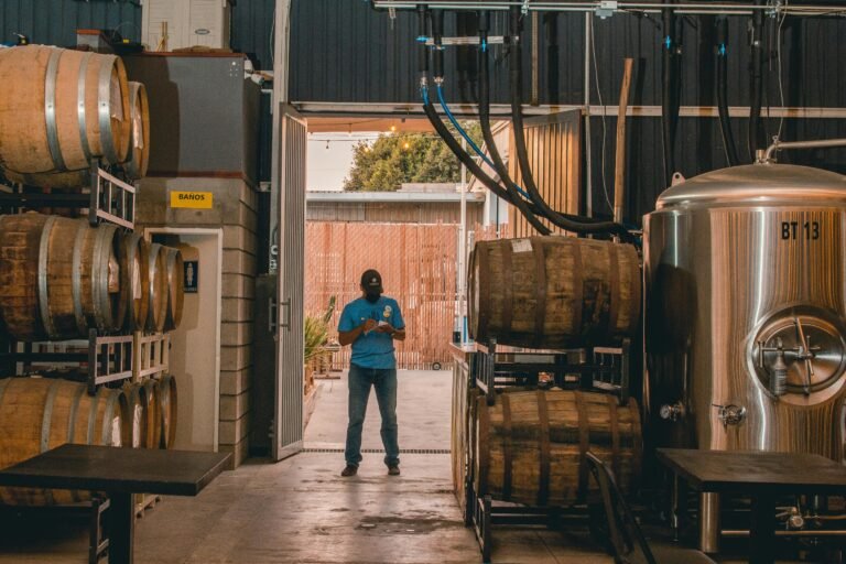 A worker stands in a brewery warehouse surrounded by kegs and equipment, creating an urban industrial scene.
